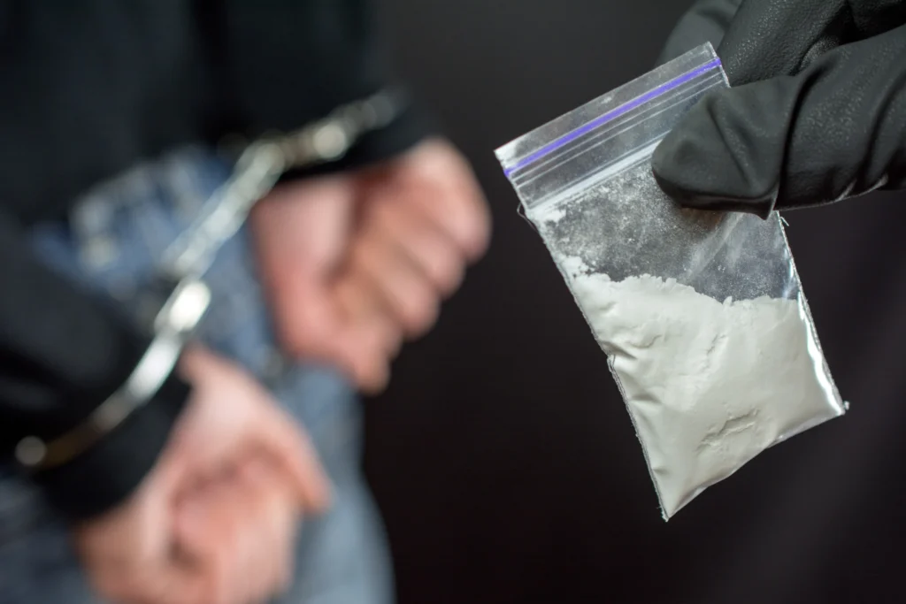 Person in handcuffs and small bag with white powder shown.