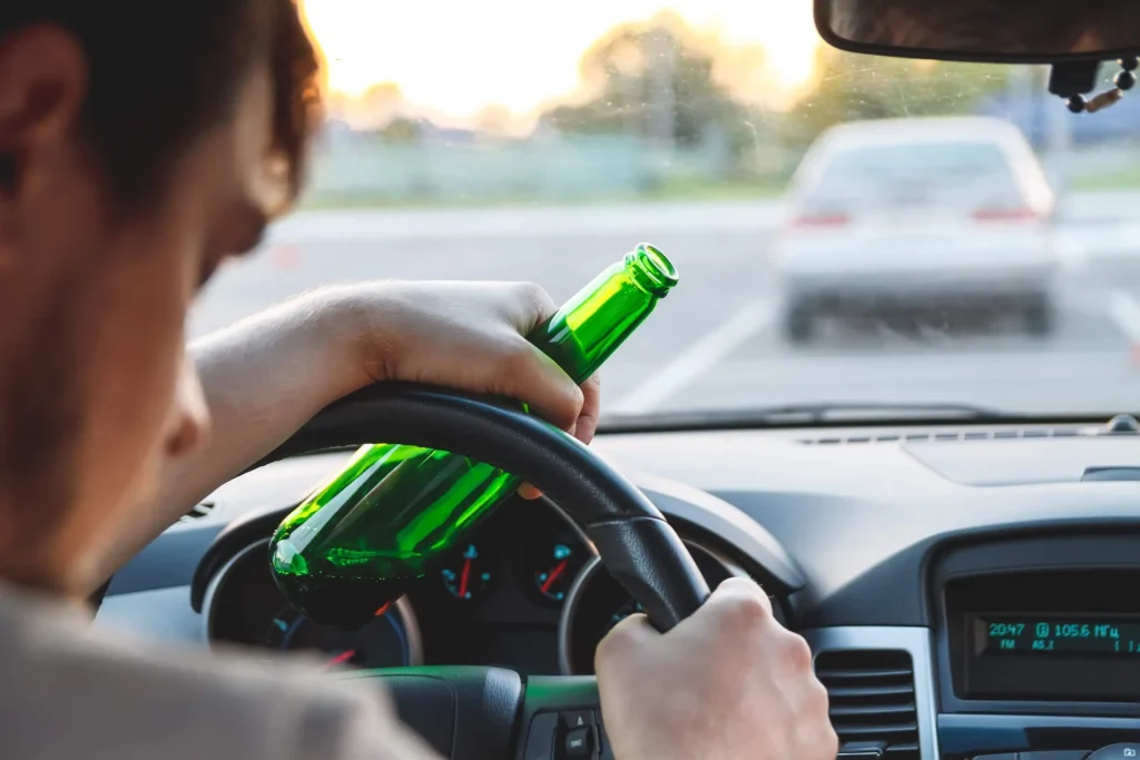Young driver behind the wheel of a car holding a beer bottle.