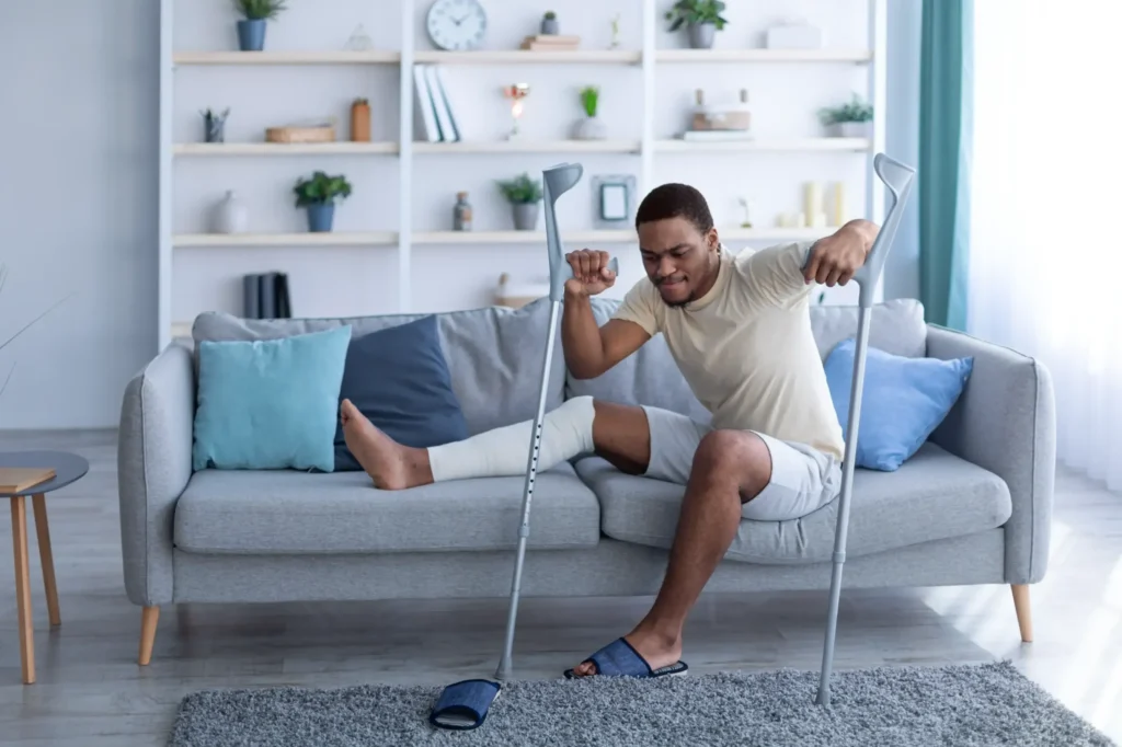 A man with a wrapped leg laying on the couch trying to use crutches to get up.